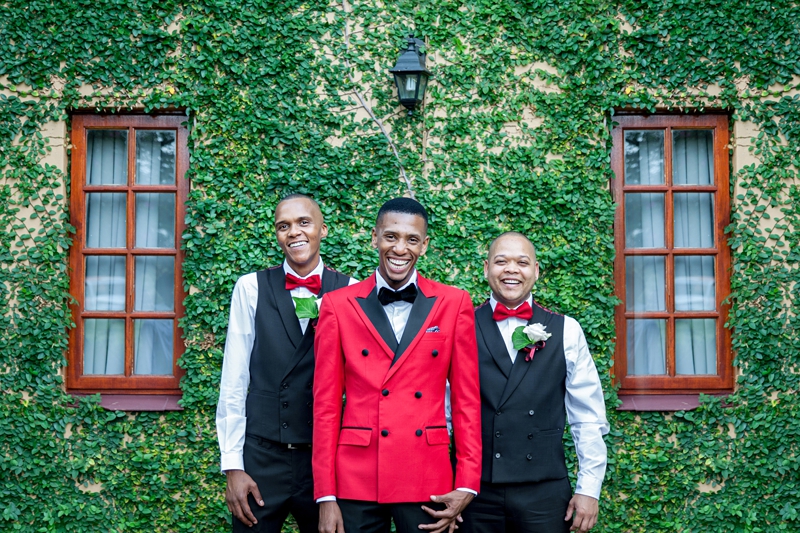 Groom, wearing red jacket, laughing with his groomsmen in front of a green wall at Valverde Eco Hotel.