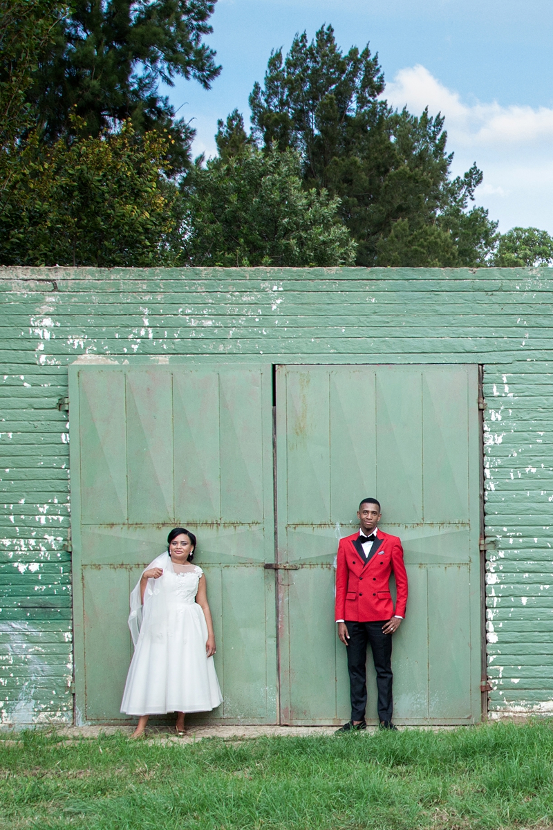 Bride and groom, wearing red jacket, standing in front of green door at Valverde Eco Hotel.