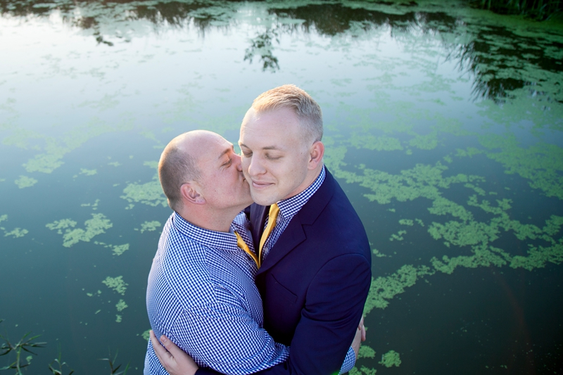 Groom kissing groom on the cheek, in front of a pond at Valverde Eco Hotel.