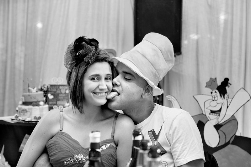 Guy, wearing large hat, licking his dates cheek during Alice in Wonderland wedding reception.