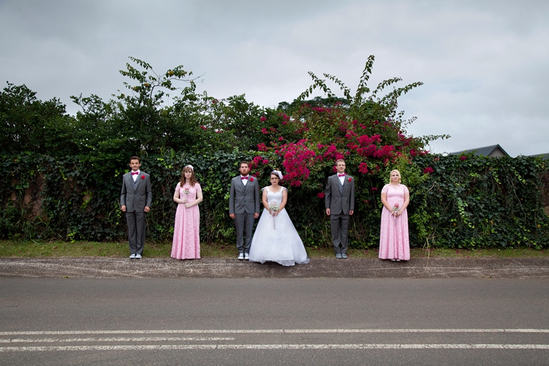 Bridal party standing on road in front of pink bougainvillea.