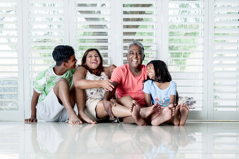 Indian family laughing while sitting on the floor, in front of shutters, in their home.