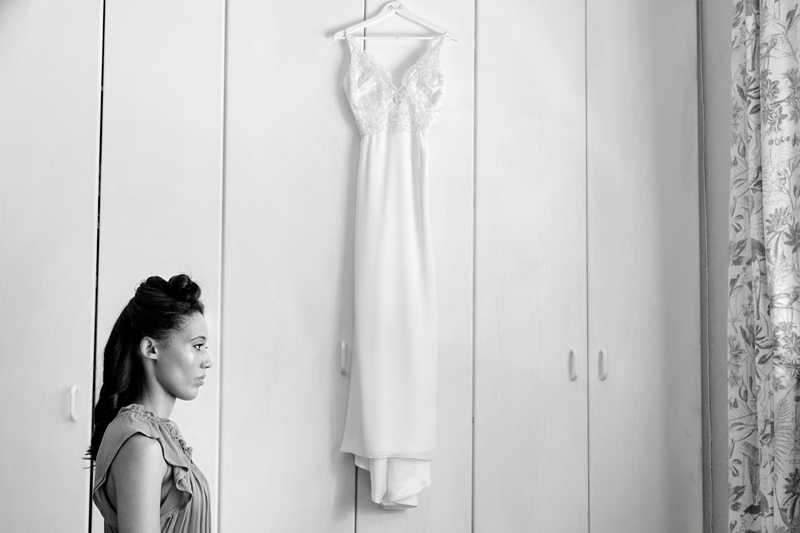 Bride getting her hair done with wedding dress hanging behind her.