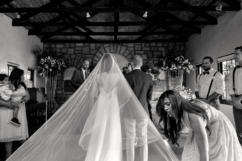 Bridesmaid fixing brides long veil during wedding ceremony at Leafy Green Cafe.