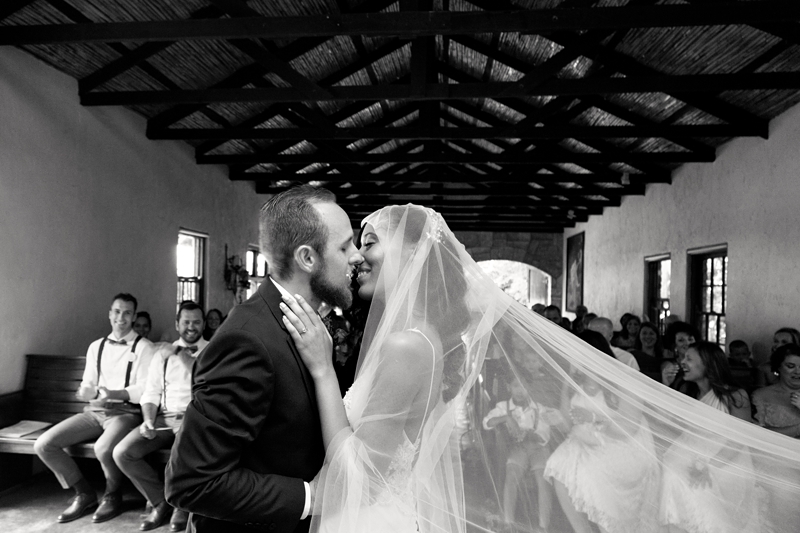 Bride, with long veil, kissing groom, with beard, during ceremony at Leafy green Cafe.