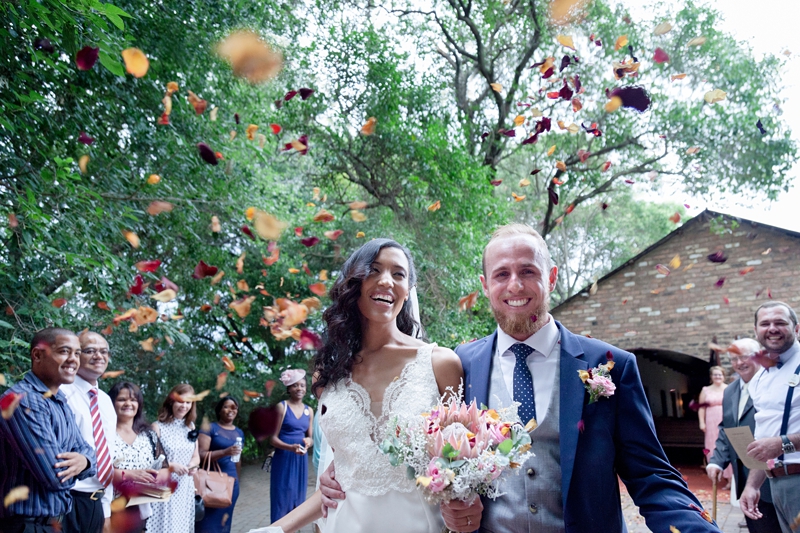 Bride and groom laughing while having red and peach rose petals thrown at them, at Leafy Green Cafe.
