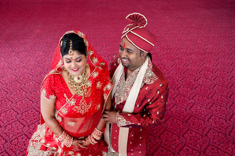 Bride and groom smiling at each other, sitting on red carpet at Shree Radhe Shyam Mandir.