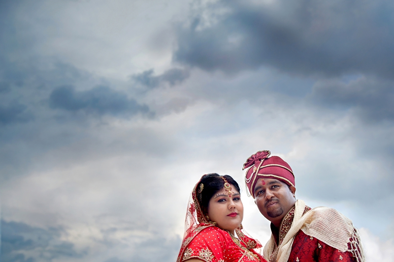 Bride and groom wearing traditional Hindu wedding outfit with clouds in the background at Shree Radhe Shyam Mandir.