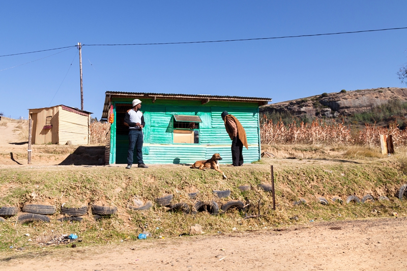 Two men and a brown dog enjoying the sun outside a turquoise Spaza shop in Lesotho.